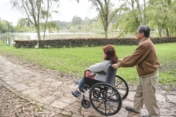 Stock image Senior hispanic couple strolling in a park, he pushes her wheelchair along a pathway.