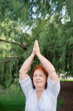 Beyaz elbiseli, kıdemli İspanyol kadın parkta yoga yapıyor. Vrksasana ayakta duruyor. Konseptler: sağlık, canlılık, aktif ve sağlıklı yaşam tarzı.