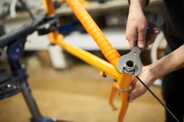 Maintenance of a bicycle: hands of an unrecognizable person using a wrench disassembling the headset of an orange bike in his workshop. Selective focus composition.