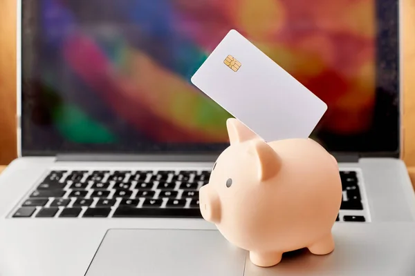 Piggy bank with a blank white credit or debit card with microchip in front of a laptop. Concepts of savings, plastic money and online shopping.