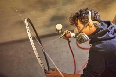 Koruyucu solunum maskesi takan İspanyol genç adam atölyesinde bisiklet kenarına sprey boya sıktı. Gerçek insanlar çalışıyor..