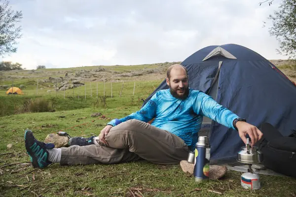 Young man sitting relaxed in a camp, in front of a tent, smiling, heating water on his stove. Concept: enjoying vacations in the open air, in contact with nature.