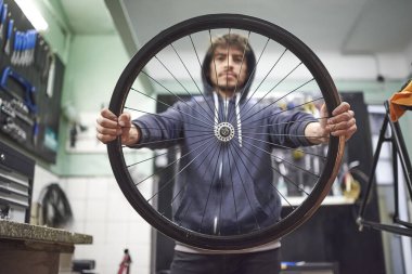 Genç İspanyol adam bisiklet tamirhanesinde bisiklet tekerleği tutuyor. Seçici odaklı kompozisyon. İş yerindeki gerçek insanlar.