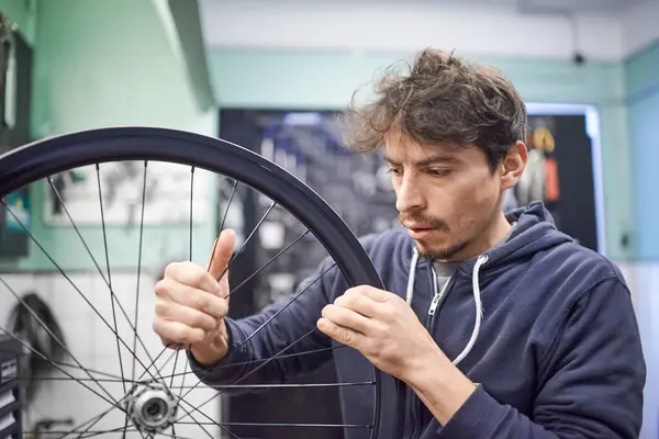 stock image Young hispanic man checking the tension of the spokes of a bicycle wheel in his bike workshop. Real people at work.