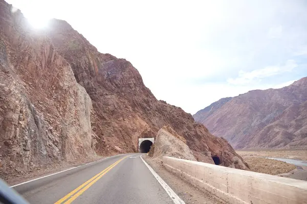 Mendoza, Arjantin 'deki yol gezisi manzarası: dağlık bir arazinin ortasında, nehrin sağında ve dağ boyunca bir tünelin önünde boş bir yol..