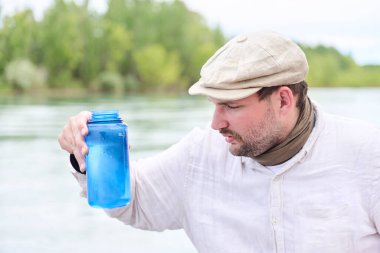 Bir adam şişesindeki nehirden su topluyor ve onun temizliğine bakıyor. Kavramları: kirlilik olmadan doğanın korunması.