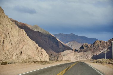 Bir araba gezisi sahnesi: Andes 'in kayalık bir dağ manzarasının ortasında boş bir yol, Arjantin' in Mendoza şehrinde, gün batımında..