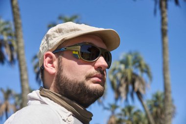 Güneşli bir günde açık havada güneş gözlüğü ve keten bereli arka planda palmiye ağaçları olan genç bir adamın portresi. El Palmar Milli Parkı 'nda doğanın tadını çıkaran bir gezgin..
