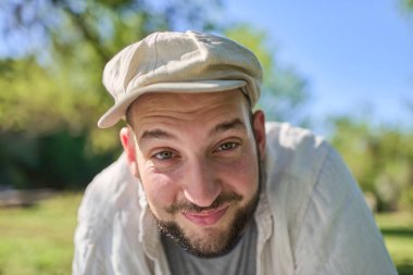 Arkadaşça gülümseyen, dışarıda iyi vakit geçiren bir adamın yakın plan portresi. Sakalı var, bere takıyor ve açık renkli gömlek giyiyor. Sahne, doğal ve sağlıklı bir ortamda eğlence ve refah taşır.