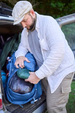 Kamp gezisi sırasında mavi sırt çantasından arabasının bagajında bir şey çıkaran bir adam..