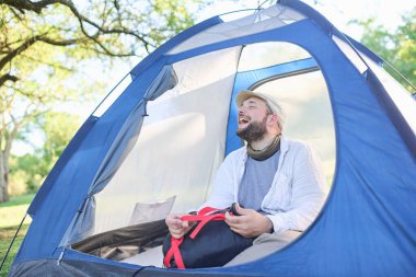 Çok heyecanlı bir genç adam, uyku tulumunu çıkarırken geniş bir gülümsemeyle vahşi bir kamp alanında çadırında oturuyor. Konsept: Seyahatin keyfi ve doğayla temasta kendini özgür hisset.