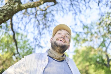 Doğada mutluluk ve refahın bir ifadesi ile gülümseyen genç bir adamın portresi; arka plan, fotokopi alanı ile gökyüzü ve odaktan çıkmış ağaçlardır. Düşük açı görünümü.