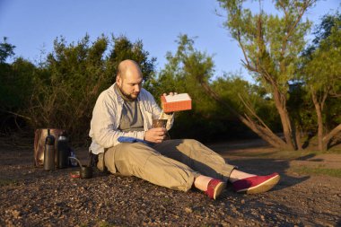 Şafak vakti bir derenin kıyısında oturan gezgin, tipik bir Arjantin içkisi olan eşi içmeye hazırlanırken ilk güneş ışınlarını alıyor. El Palmar Ulusal Parkı, Entre Rios, Arjantin.