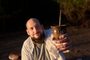 Gülümseyen adam kameraya eş teklif ediyor. Bu Arjantin içkisi paylaşımın ve birlikteliğin sembolüdür. Seçici bir şekilde ele odaklı kompozisyon.