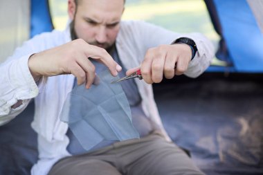 Bir çadırda oturan erkek gezgin çadırındaki deliği onarmak için çok fonksiyonlu aletinin makası ile sivrisinek ağı kesiyor..