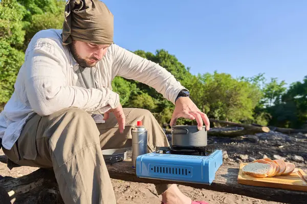 Açık mavi portatif bir ocakta güneşli bir sabah kamp alanında kamp yaparken İspanyol bir adam yemek pişiriyor..