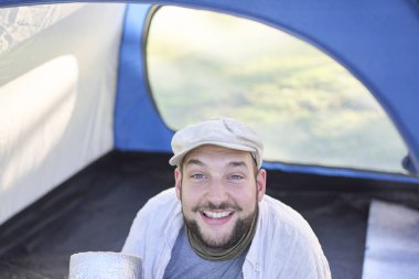 Mutlu gezgin kameraya bakıyor ve vahşi doğada kamp yaparken çadırında oturmuş gülümsüyor. Konsept: seyahat etmenin ve doğayla temasta özgür hissetmenin mutluluğu ve refahı.