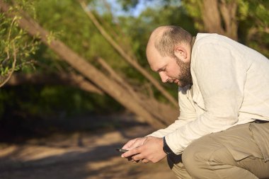 Adam dışarıda oturuyor, doğada, sosyal medya hesaplarını kontrol etmek için akıllı telefonunu kullanıyor..