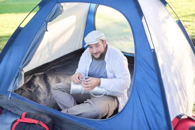 Vahşi doğada kamp yaparken çadırında oturan mutlu gezgin kameraya bakıp gülümsüyor. Konsept: seyahat etmenin ve doğayla temasta özgür hissetmenin mutluluğu ve refahı.
