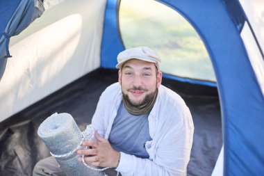 Mutlu gezgin kameraya bakıyor ve vahşi doğada kamp yaparken çadırında oturmuş gülümsüyor. Konsept: seyahat etmenin ve doğayla temasta özgür hissetmenin mutluluğu ve refahı.