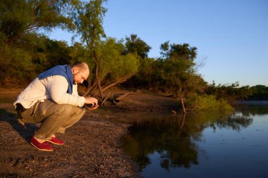Gezgin şafak vakti, güneşin ilk ışıklarıyla aydınlatılan sakin doğal bir manzarada ellerini ve yüzünü bir derenin kıyısında yıkıyor. El Palmar Ulusal Parkı, Entre Rios, Arjantin.