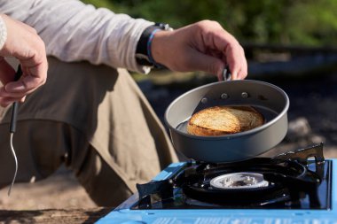 Tanınmayan biri, sabah kamp alanında kahvaltıda, gaz ocağında tavayla ekmek kızartıyor..