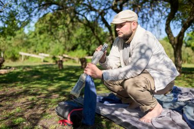 Genç adam çadır direklerini elinde tutuyor ve onu barışçıl bir kamp alanına kurma sürecini başlatıyor. Çevre yeşil ve hava güneşli..