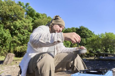 İspanyol gezgin, güneşli bir sabah kamp alanında kamp yaparken açık mavi portatif bir fırında yemek pişiriyor..