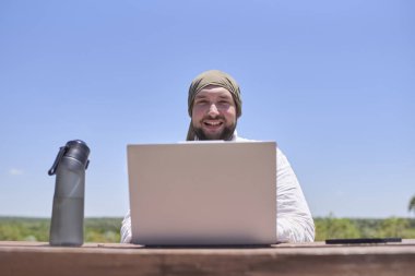 Güneşli bir günde açık hava masasında dizüstü bilgisayarıyla çalışırken gülümseyen genç bir İspanyol. Dijital göçebe, özgürlüğün tadını çıkarırken uzaktan çalışıyor..