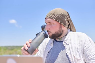 Güneşli bir günde laptopuyla dışarıda çalışırken tekrar kullanılabilir bir şişeden su içen genç bir İspanyol. Konsept: Seyahat ederken uzaktan çalışan dijital göçebenin sağlıklı yaşam tarzı.