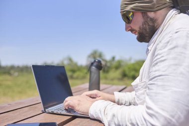 Güneşli bir günde dışarıda bir masada dizüstü bilgisayarıyla çalışan genç bir İspanyol. Dijital göçebe, özgürlüğün tadını çıkarırken uzaktan çalışıyor..