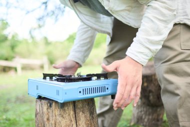 Doğa kampında kamp yaparken açık mavi portatif bir fırının üzerinde yemek pişirmek üzere olan tanınmayan biri..