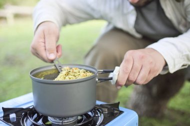 Tanınmayan kişi dışarıda sebzeli yemek pişiriyor sebzeli pilav yapıyor, taşınabilir gaz sobası kullanıyor ve kamp yapıyor. Bir el tencereyi tutarken diğer el kaşıkla yemeği karıştırıyor..