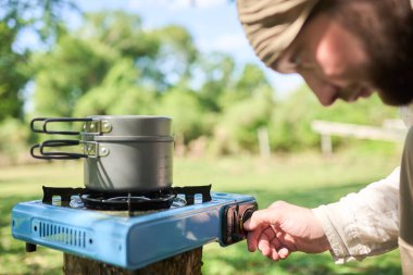 Erkek gezgin doğa kampında kamp yaparken dışarıda yemek pişiriyor güneşli bir günde, üzerinde bir tencere ile taşınabilir bir gaz sobası yakıyor..