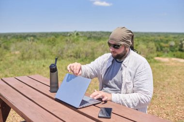 Güneşli bir günde dışarıda bir masada dizüstü bilgisayarıyla çalışan genç bir İspanyol. Dijital göçebe, özgürlüğün tadını çıkarırken uzaktan çalışıyor..