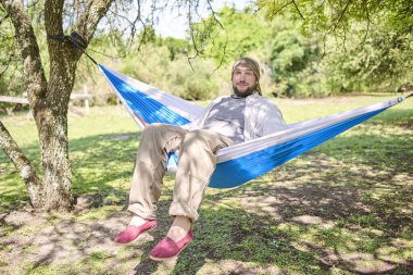 Bir hamakta otururken kameraya gülümseyen ve dışarıda huzur içinde dinlenen genç bir adamın portresi..