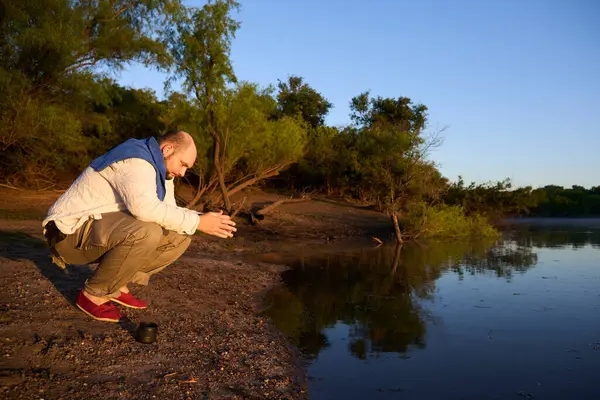 Gezgin, güneşin ilk ışıklarıyla aydınlatılan sakin doğal bir manzarada gün doğumunda bir derenin kıyısında ellerini ve yüzünü yıkıyor. El Palmar Ulusal Parkı, Entre Rios, Arjantin.