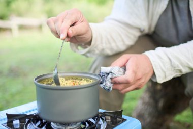 Tanınmayan kişi dışarıda sebzeli yemek pişiriyor sebzeli pilav yapıyor, taşınabilir gaz sobası kullanıyor ve kamp yapıyor. Bir el tencereyi tutarken diğer el kaşıkla yemeği karıştırıyor..