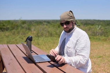 Genç bir İspanyol, güneşli bir günde açık hava masasında dizüstü bilgisayarıyla çalışırken telefonunu kontrol ediyor. Dijital göçebe, özgürlüğün tadını çıkarırken uzaktan çalışıyor..