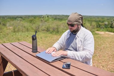 Genç İspanyol adam uzaktan kumandalı işini bitirdikten sonra bilgisayarını kapatıyor, açık havada güneşli bir doğal ortamda. Dijital göçebe seyahat ederken çalışıyor, özgürlüğün tadını çıkarıyor..