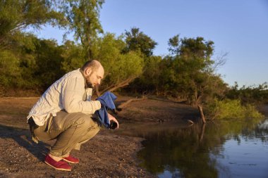 İspanyol gezgin, güneşin ilk ışıklarıyla aydınlanan sakin doğal bir manzarada, gün doğumunda ellerini ve yüzünü bir derenin kıyısında yıkıyor. El Palmar Ulusal Parkı, Entre Rios, Arjantin.