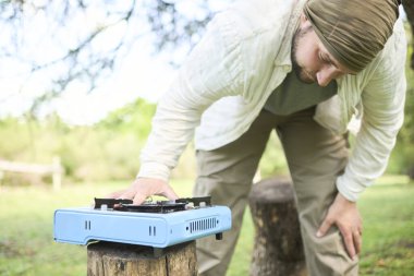 Genç İspanyol bir adam doğa kampında kamp yaparken açık mavi portatif bir ocakta yemek pişirmek üzere..