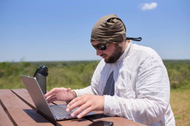 Güneşli bir günde dışarıda bir masada dizüstü bilgisayarıyla çalışan genç bir İspanyol. Dijital göçebe, özgürlüğün tadını çıkarırken uzaktan çalışıyor..