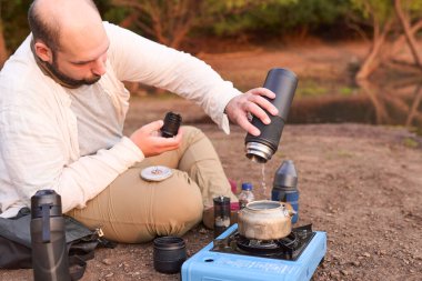 İspanyol bir adam, gün doğarken bir derenin kıyısında ısıtmak için çaydanlığa su döküyor. El Palmar Ulusal Parkı, Entre Rios, Arjantin.