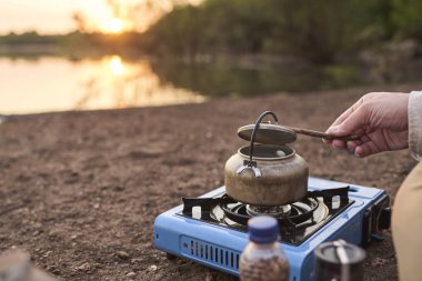 Tanınmayan biri, gün doğumunda kahve yapmak için çaydanlıkta su ısıtırken bir derenin kenarında bir seyahat sobasıyla su ısıtıyor. El Palmar Ulusal Parkı, Entre Rios, Arjantin.
