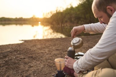 Şafak vakti, bir çaydanlıktan termosa sıcak su döken kişi, açık havada, bir derenin kıyısında. Kavram: Doğadaki huzur ve güzelliğin özel anlarından zevk almak için seyahat etmek. Entre Rios, Arjantin.