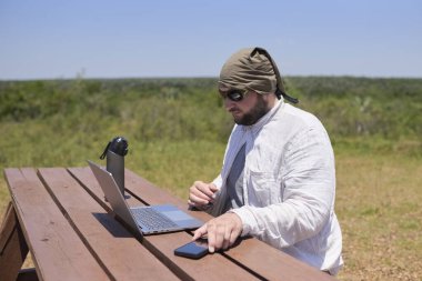 Güneşli bir günde dışarıda bir masada dizüstü bilgisayarıyla çalışan genç bir İspanyol. Dijital göçebe, özgürlüğün tadını çıkarırken uzaktan çalışıyor..