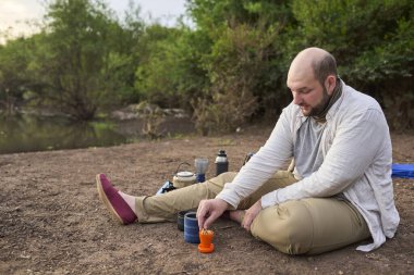 İspanyol bir adam kahvaltı yapıyor, gün doğumunda bir derenin kıyısında taze demlenmiş kurabiyelerle bir fincan kahvenin tadını çıkarıyor. El Palmar Ulusal Parkı, Entre Rios, Arjantin.