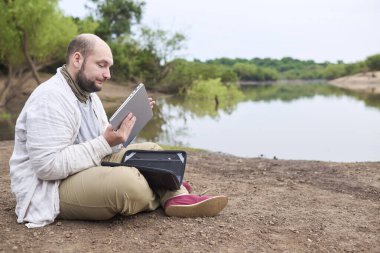 Adam bir süre akarsu kenarında çalışmak için bilgisayarını çıkarıyor. Konsept: dijital göçebe seyahat ederken uzaktan çalışıyor, özgürlüğün tadını çıkarıyor.