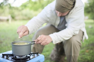 Genç adam dışarıda vejetaryen yemeği pişiriyor, sebzeli pilav pişiriyor, açık mavi taşınabilir gaz ocağında pişirme kabı kullanıyor, doğa kampında kamp kuruyor. Esrara odaklan..